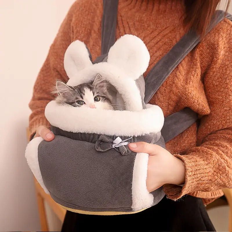 tabby and white fluffy kitten, sits in a grey and white carrier bag, strapped onto a woman, wearing a brown pullover. 