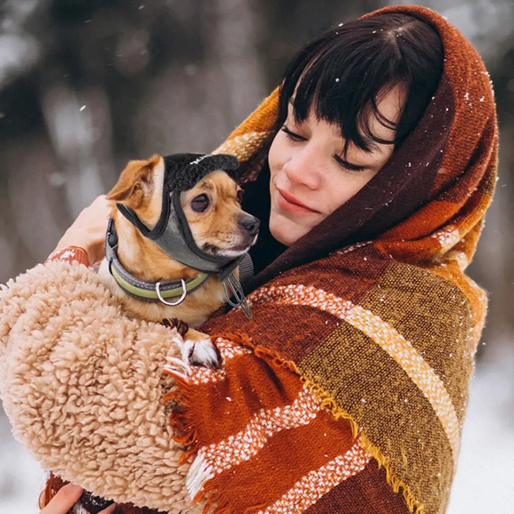 a woman holds a small dog wearing a black winter dog hat, in a snowy setting. 