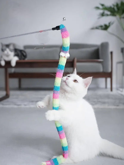 White kitten plays with a Purrfect Pounce Teaser Wand in a home setting, another cat lies on a table in the background, watching. 