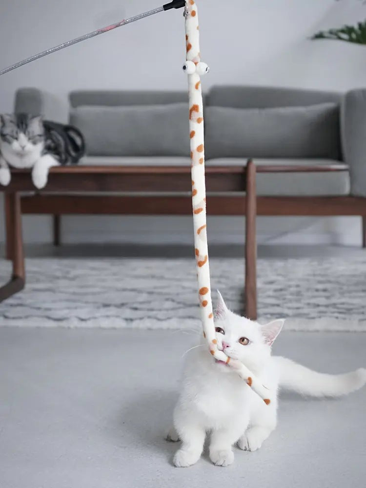 White kitten plays with a Purrfect Pounce Teaser Wand in a home setting, another cat lies on a table in the background, watching. 