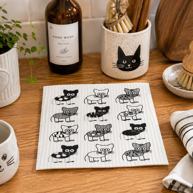 A square kitchen towel with a white ribbed background and a black-and-gray cartoon cat pattern, displayed on a wooden countertop in a styled kitchen scene.