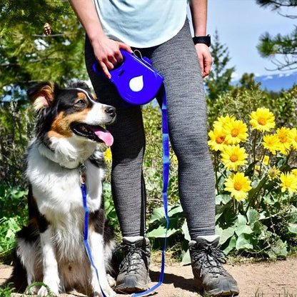 Outdoor setting, blue sky, trees, yellow flowers. A dog sits by a person's side, who is holding a blue leash, attached to the dog's collar. 